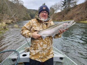 Angler in camouflage jacket holding steelhead on small boat in river with forested hillsides