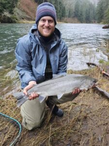 Angler kneeling on riverbank holding fresh steelhead with forested river and mountains in background