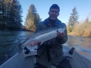 Angler in drift boat holding steelhead on forested river in winter conditions