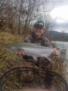 Angler in camouflage waders holding large steelhead by river with bare trees and mountains in background.