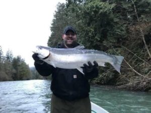 Angler in black jacket holding large steelhead on boat in green river surrounded by trees