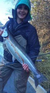 Angler in blue beanie and dark jacket displaying large steelhead trout at winter riverbank location.