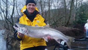 Angler in yellow jacket holding large steelhead at riverbank during winter fishing trip