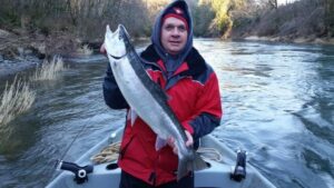 Angler in red jacket holding large steelhead while standing in drift boat on forested river