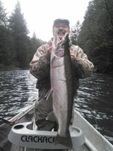 Angler in camouflage jacket holding large steelhead in small boat on forested river under overcast sky.