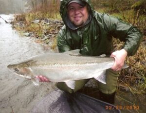 Angler in green rain jacket holding large steelhead by riverbank in rainy conditions