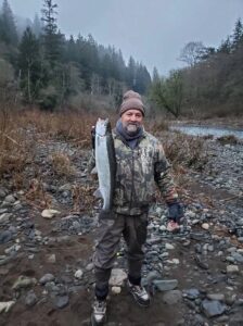 Angler in camouflage jacket holding steelhead on rocky riverbank with forested hills in background
