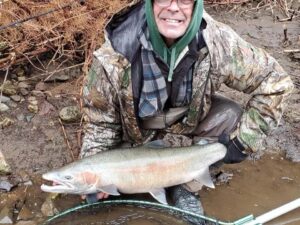 Angler in camouflage jacket kneeling with large steelhead on muddy riverbank beside fishing net.