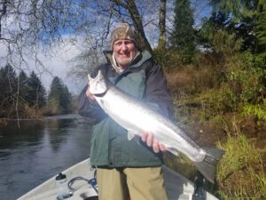 Angler in boat holding large silver steelhead on winter river surrounded by trees and clear sky