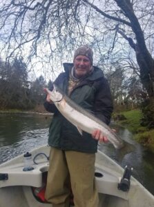 Angler in camouflage hat holding large steelhead in small boat on overcast winter river day