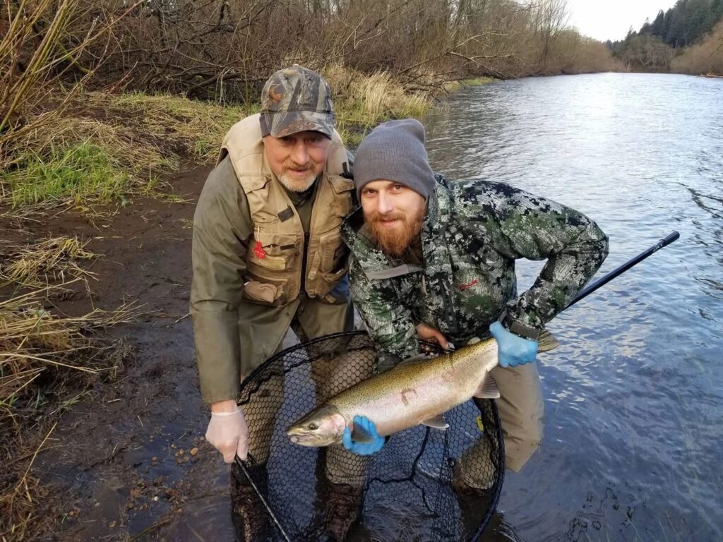Two anglers holding a large steelhead in landing net at muddy riverbank with bare winter vegetation.
