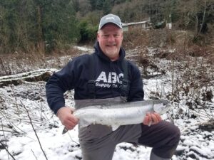 Angler kneeling in snow holding large steelhead on winter riverbank with house visible in background