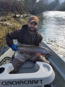 Angler in inflatable pontoon boat holding steelhead on winter river with bare trees along bank.
