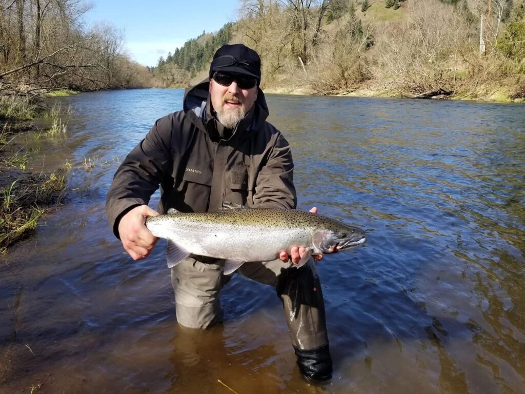 Angler in waders holding large steelhead while standing in shallow river with forested hills behind