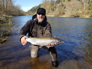 Angler in waders holding large steelhead while standing in shallow river with forested hills behind