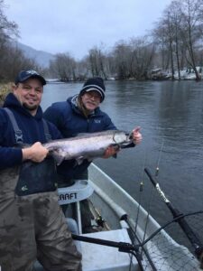 Two anglers in drift boat holding large steelhead on winter river with bare trees and snow.
