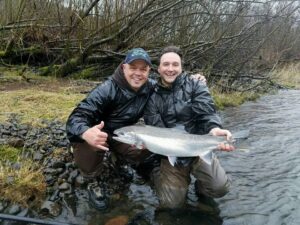 Two anglers kneeling in shallow water holding a large steelhead on Oregon riverbank