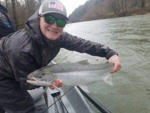 Angler in camouflage jacket holding large steelhead in drift boat on overcast river day