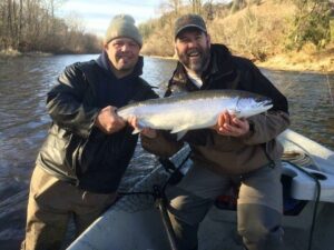 Two anglers in winter gear holding large steelhead on drift boat in river setting