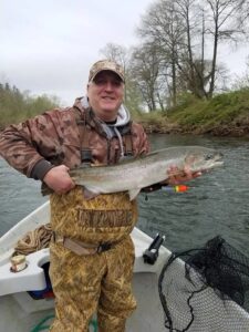 Angler in camouflage waders holds steelhead in drift boat on overcast river with bare trees