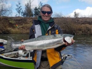 Angler in sunglasses and jacket holding large steelhead in drift boat on river with hillside background