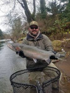 Angler holding large steelhead by river bank with landing net and bare trees in background