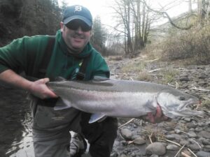 Angler in green hoodie and Detroit cap holding large steelhead on rocky riverbank in winter