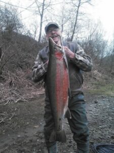 Angler holding large steelhead at riverbank during winter with bare trees in background