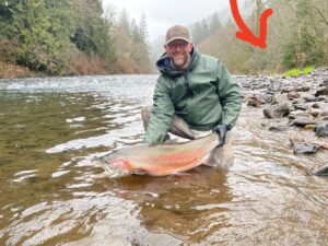 Angler kneeling in shallow river water holding large steelhead with pink stripe on rocky shoreline.