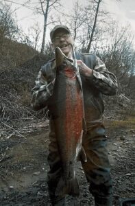 Angler holding large steelhead trout at riverbank with bare trees in background during winter