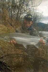 Angler in camouflage gear kneeling on riverbank holding large steelhead with bare winter trees behind