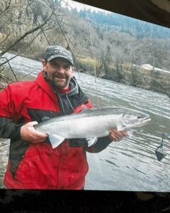 Angler in red jacket holding steelhead on river with forested hillside in background