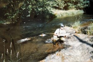 Angler in white shirt and cap displaying salmon while kneeling on riverside rocks with flowing water and vegetation behind