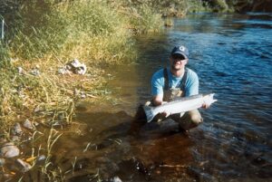 Angler kneeling in shallow river water holding a long silver salmon near reeds and grasses.
