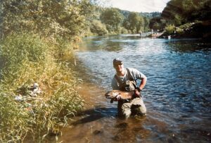 Angler kneeling in shallow river water displaying a salmon catch with forested hills in background