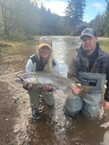 Two anglers in waders holding large salmon in shallow river water with forested hills behind