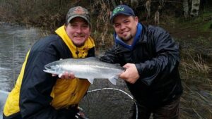 Two anglers holding a silver salmon at wooded riverbank with fishing net visible.