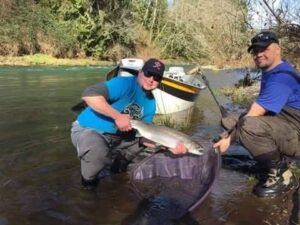 Two anglers kneeling in shallow river water displaying a large salmon in a landing net with drift boat behind.