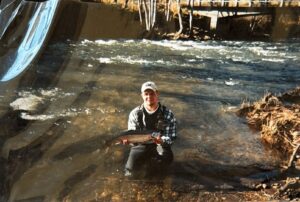 Angler kneeling in shallow river holding a salmon under a bridge with flowing water behind