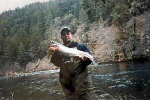 Angler in waders holding silver salmon in shallow river with forested hillside background