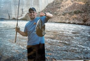 Angler in blue shirt holding caught salmon while standing in shallow river water near rocky bank
