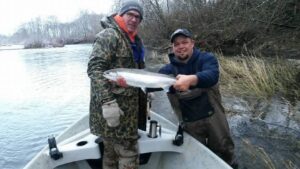 Two anglers in boat holding large silver salmon on winter river with bare trees along shoreline