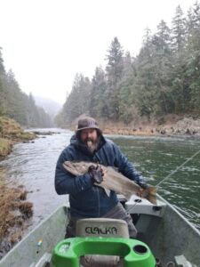 Bearded angler in winter jacket holding large salmonid fish from small boat on forested river.