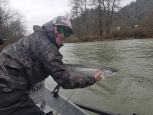Angler in camouflage jacket holding a salmon while seated in an aluminum boat on a river.
