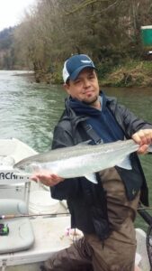 Angler holding large silvery salmon on boat in river with forested banks and greenish water