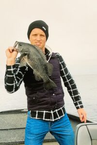 Angler in black beanie and plaid shirt holding a dark-colored fish on a boat in foggy conditions.