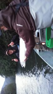 Person holding a silver salmon on a boat with water and shoreline visible in background.