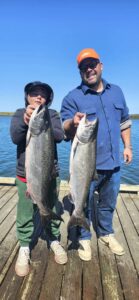 Adult and child holding two large chinook salmon on wooden dock with blue water and clear sky behind them