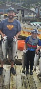 Father and son holding four chinook salmon on wooden dock with yellow fishing boat and coastal buildings behind them.