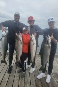 Family of four displaying four large chinook salmon on wooden dock under overcast sky with water and mountains behind